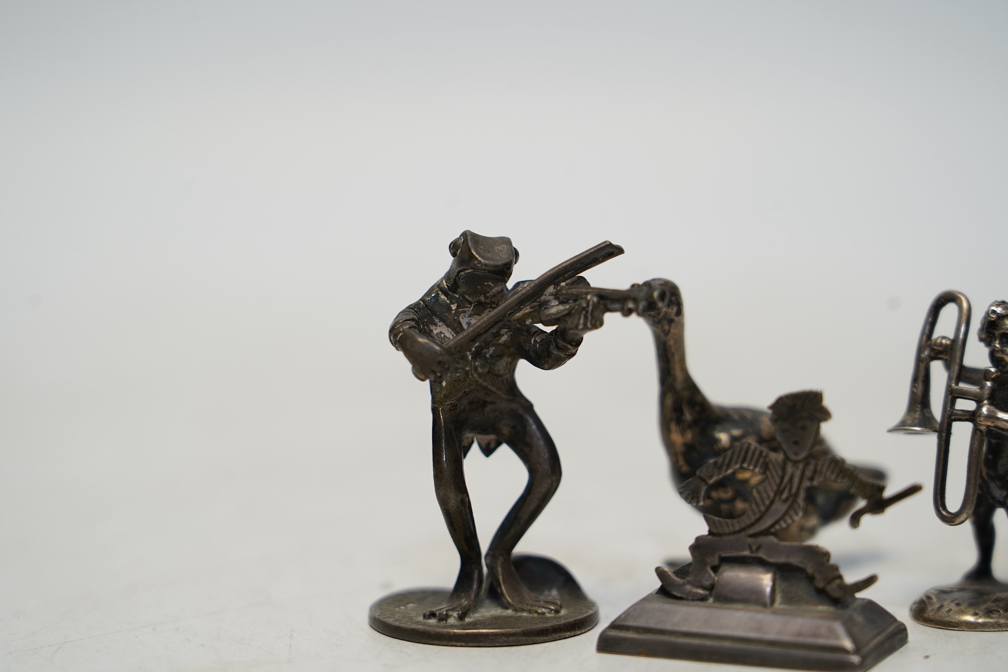 Two early 20th century miniature silver models of a goose and a cherub with trombone, 37mm, an 800 standard figural menu holder and a later silver model of a frog with violin. Condition - fair